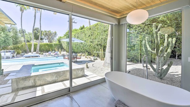 A modern bathroom with a freestanding bathtub, large windows, and a view of a pool and desert landscape.