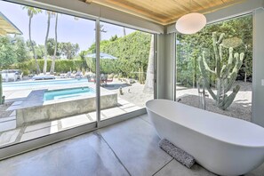 Um banheiro moderno com uma banheira separada, grandes janelas e vista para uma piscina e paisagem desértica.