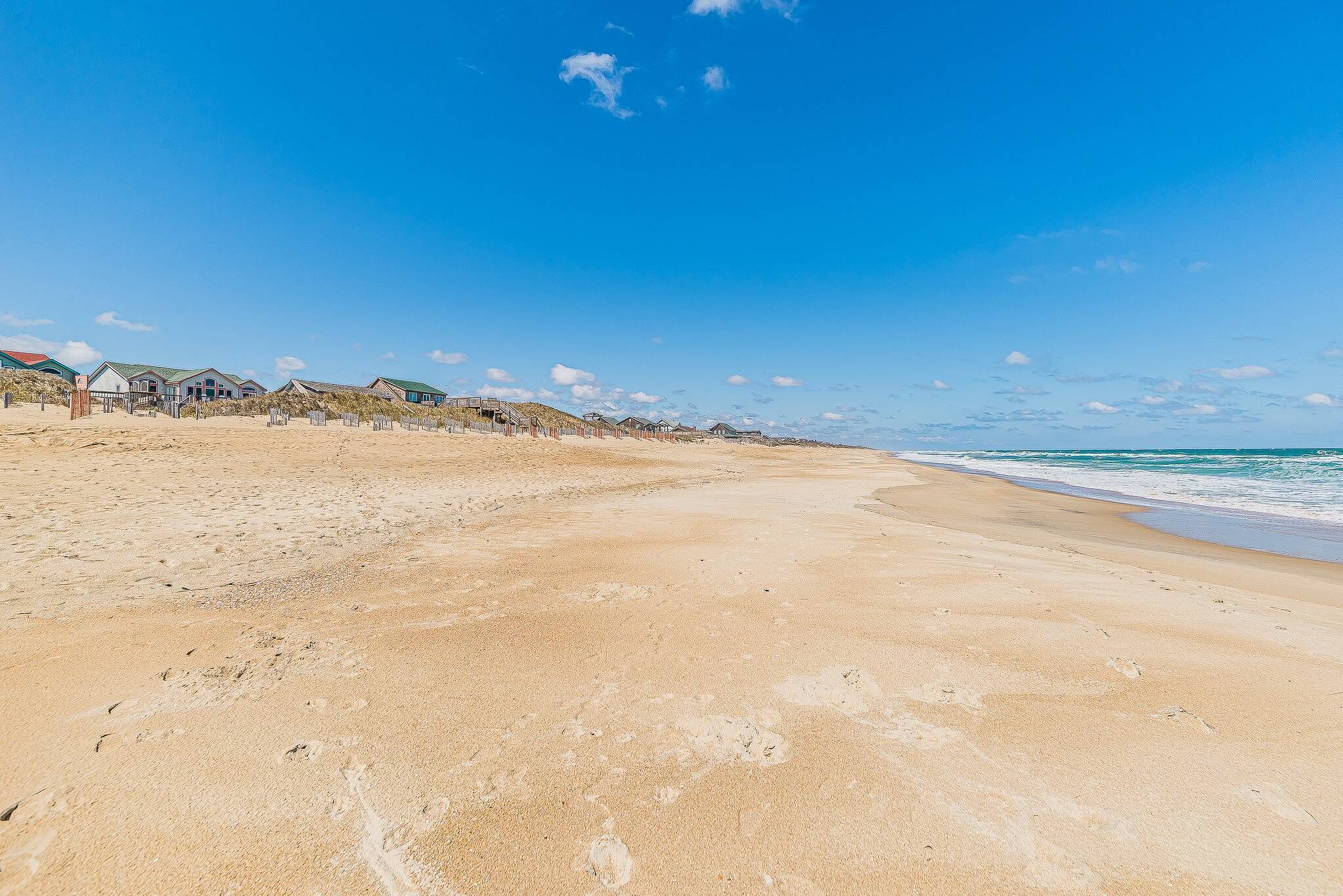 A sandy beach with footprints, a clear blue sky, and beach houses in the background.