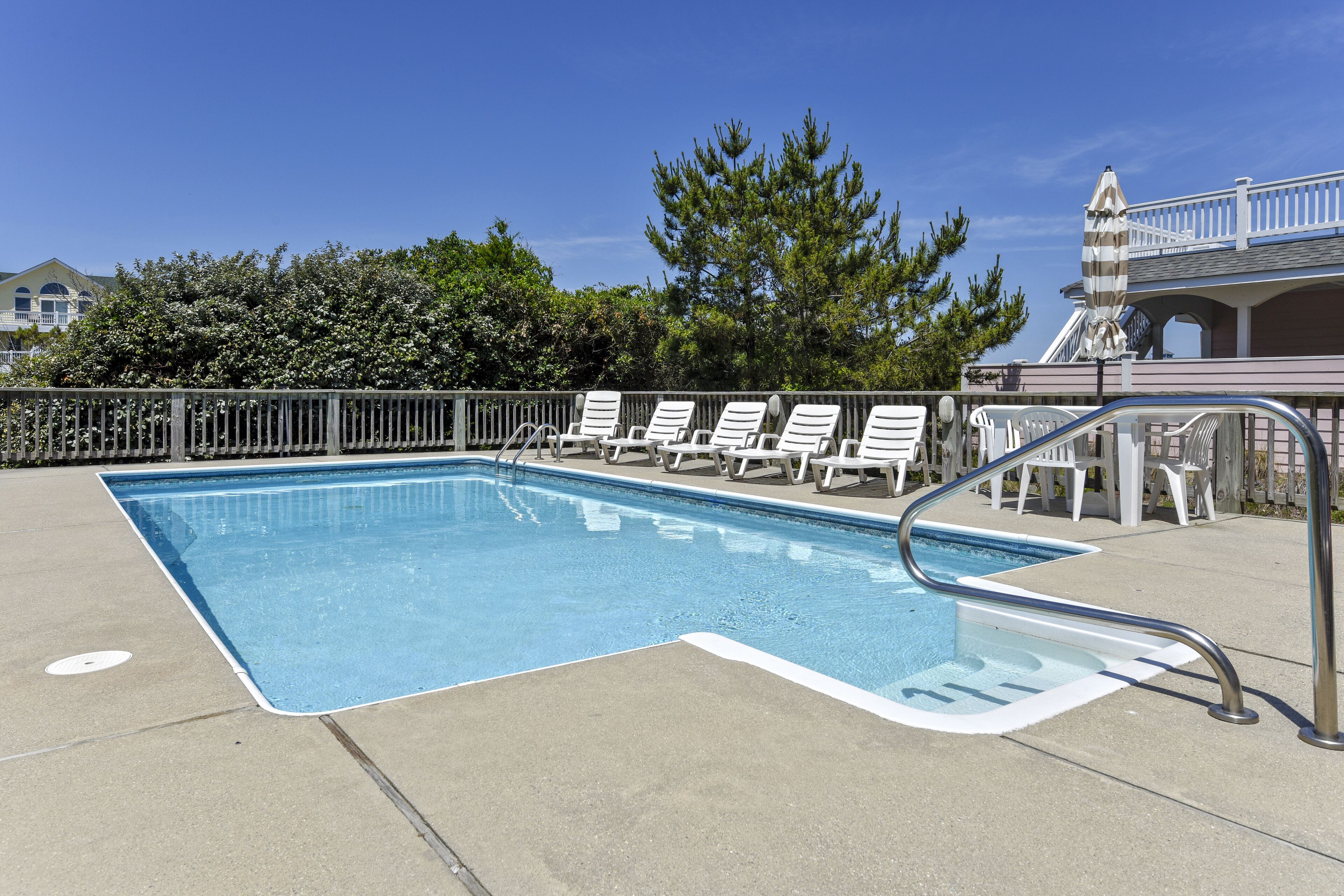 A rectangular pool with a metal ladder, surrounded by white lounge chairs and a wooden fence.