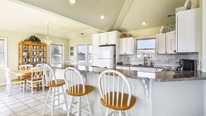 A modern kitchen with white cabinets, a granite countertop, and a dining area with white chairs and a wooden table. - WH816- Remarkable! 6 BRs, Private Pool, Hot Tub and even a Volleyball Court! (Corolla)