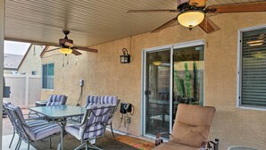 Un patio exterior con una mesa y sillas, un ventilador de techo y un ventanal corredizo que da a un jardín.