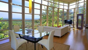 Ein modernes Wohnzimmer mit Glastisch, weißen Stühlen, einer Couch und einem Fernseher. Große Fenster bieten Blick auf Grünflächen und eine entfernte Landschaft.