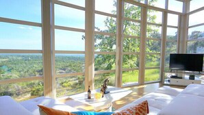 Ein geräumiges Zimmer mit großen Fenstern und Blick auf eine üppige Landschaft. Es gibt ein weißes Sofa mit orangefarbenen und grünen Kissen, einen kleinen Tisch mit einem dekorativen Gegenstand sowie einen Flachbildfernseher auf einem Standfuß.