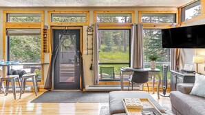 Ein geräumiger Wohnbereich mit einer großen Couch, einem Couchtisch und einem Fernseher. Mehrere Fenster sorgen für natürliches Licht und Ausblicke ins Freie.