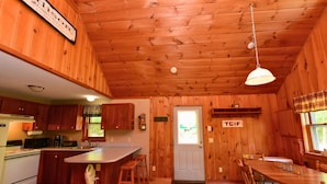 Interior de una cabaña con paredes de madera, una cocina con estufa, una zona de comedor con mesa y sillas, y una puerta con ventana.