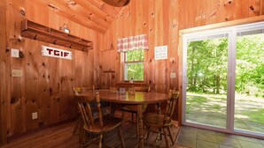 Interior de una cabaña de madera con una mesa redonda, cuatro sillas de madera, una ventana con cortina a cuadros y un cartel que dice 'Life is better at the cabin'.