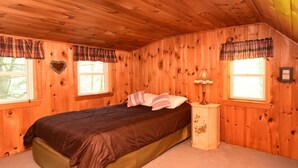 Un dormitorio de cabaña de madera con cama, dos ventanas, un tocador y cortinas a cuadros.