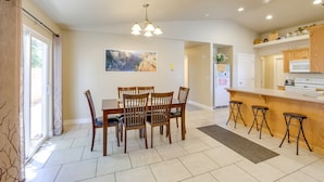 A dining area with a table and chairs, a kitchen with a counter and stools, and a large window with curtains. - Family-Friendly Utah Home, Easy Access to Zion! (La Verkin)