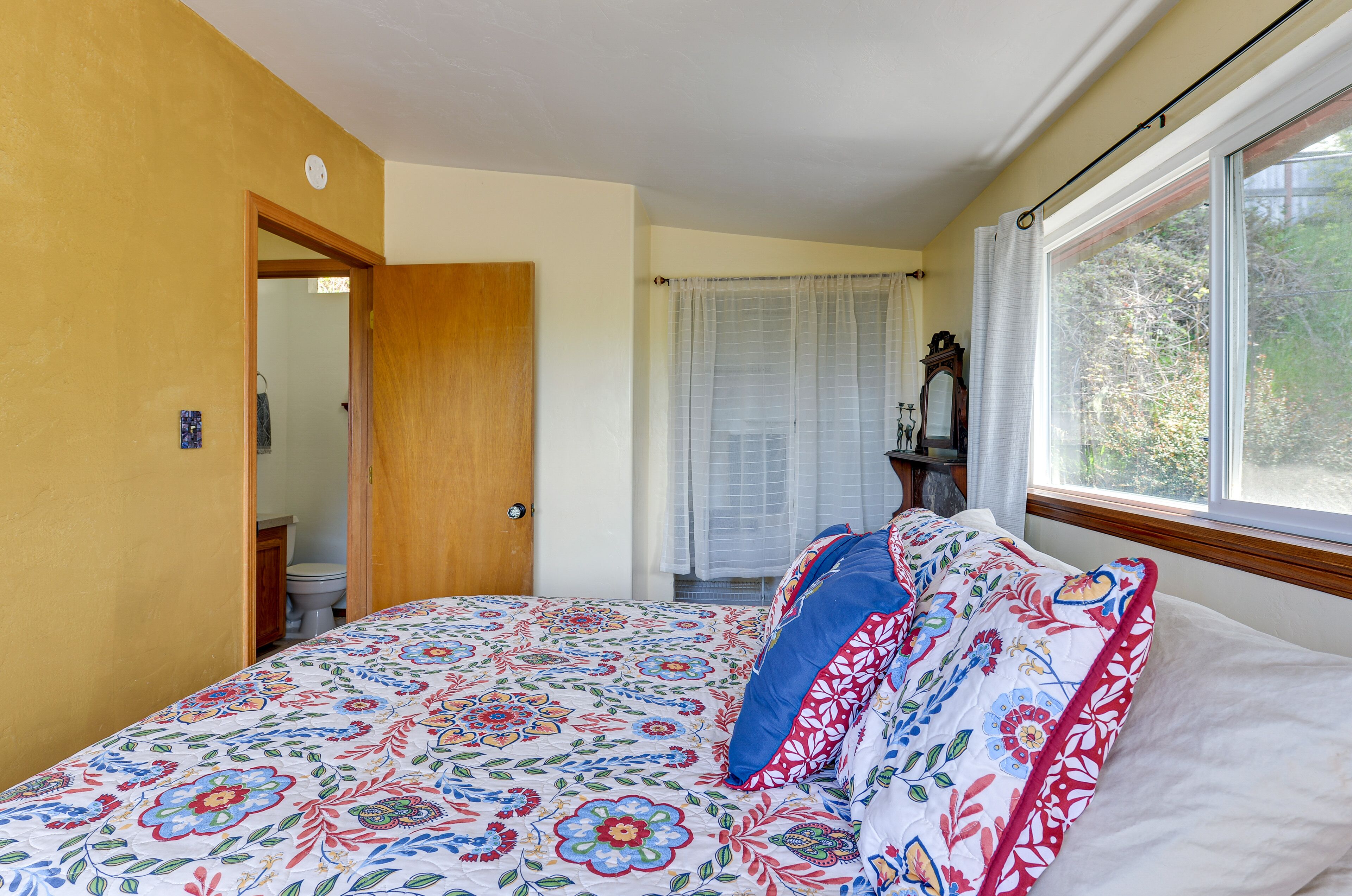 A bedroom with a floral bedspread, a wooden door, a window with curtains, and a view of trees outside.