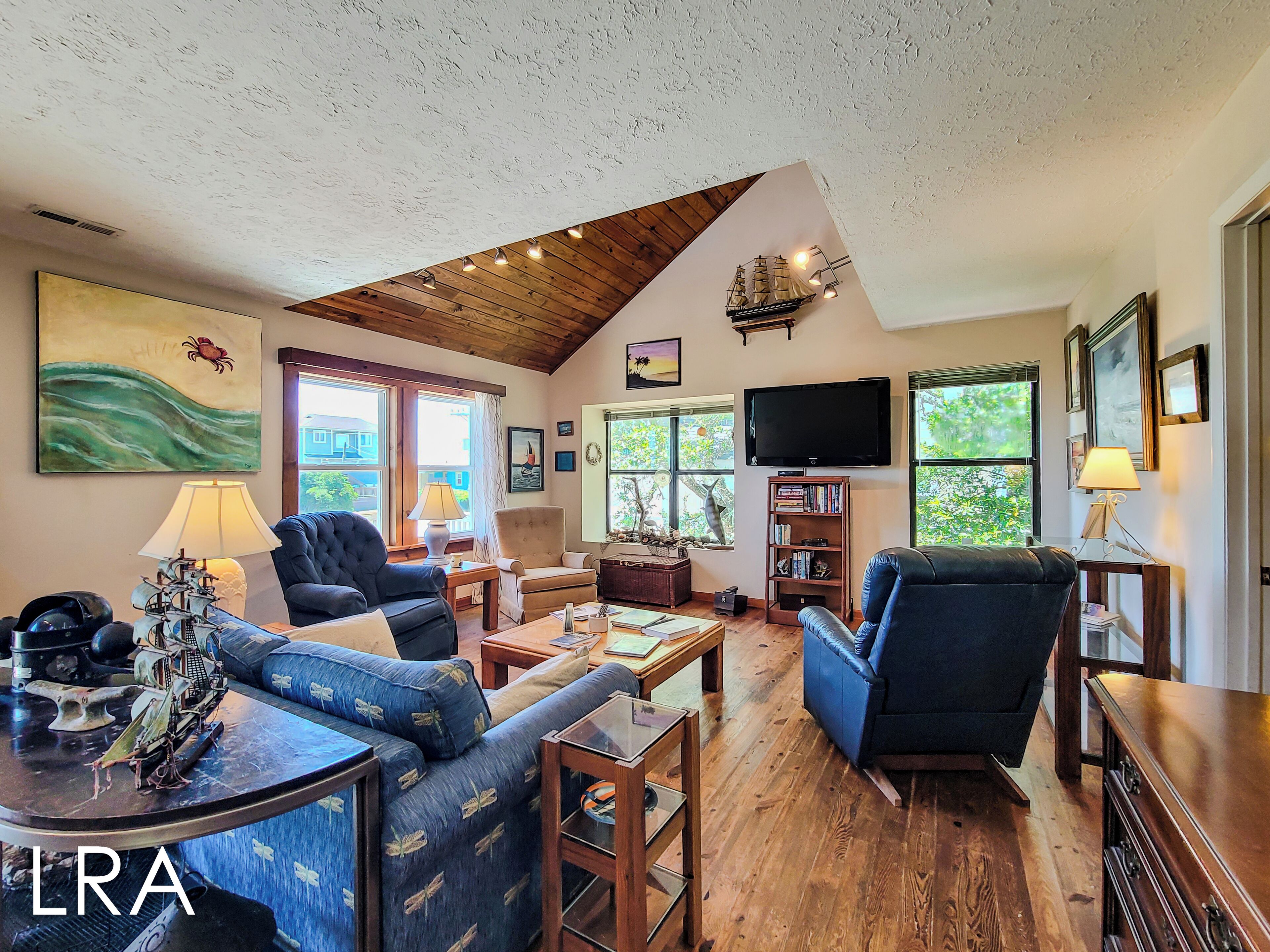 A spacious living room with a high ceiling, wooden flooring, and a large window offering a view of greenery outside.