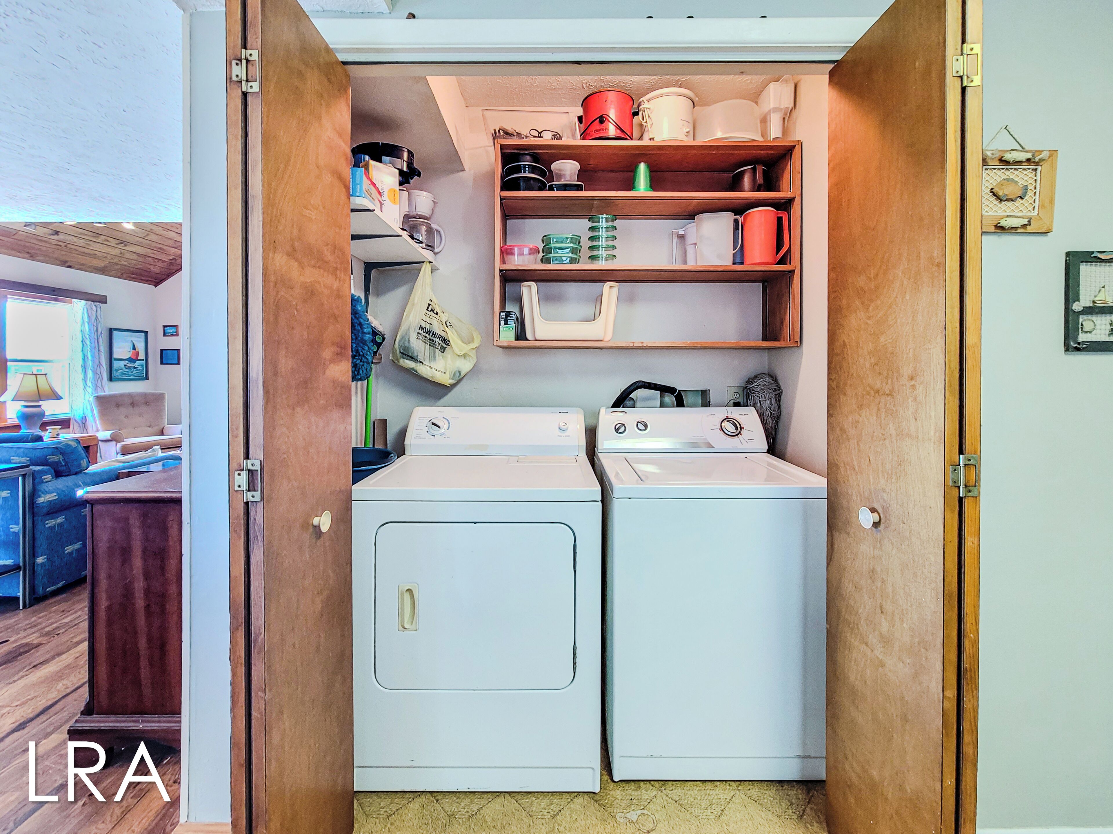 A laundry room with two washing machines, shelves above containing various containers and a bag, and a door leading to a living area with a sofa and a lamp.