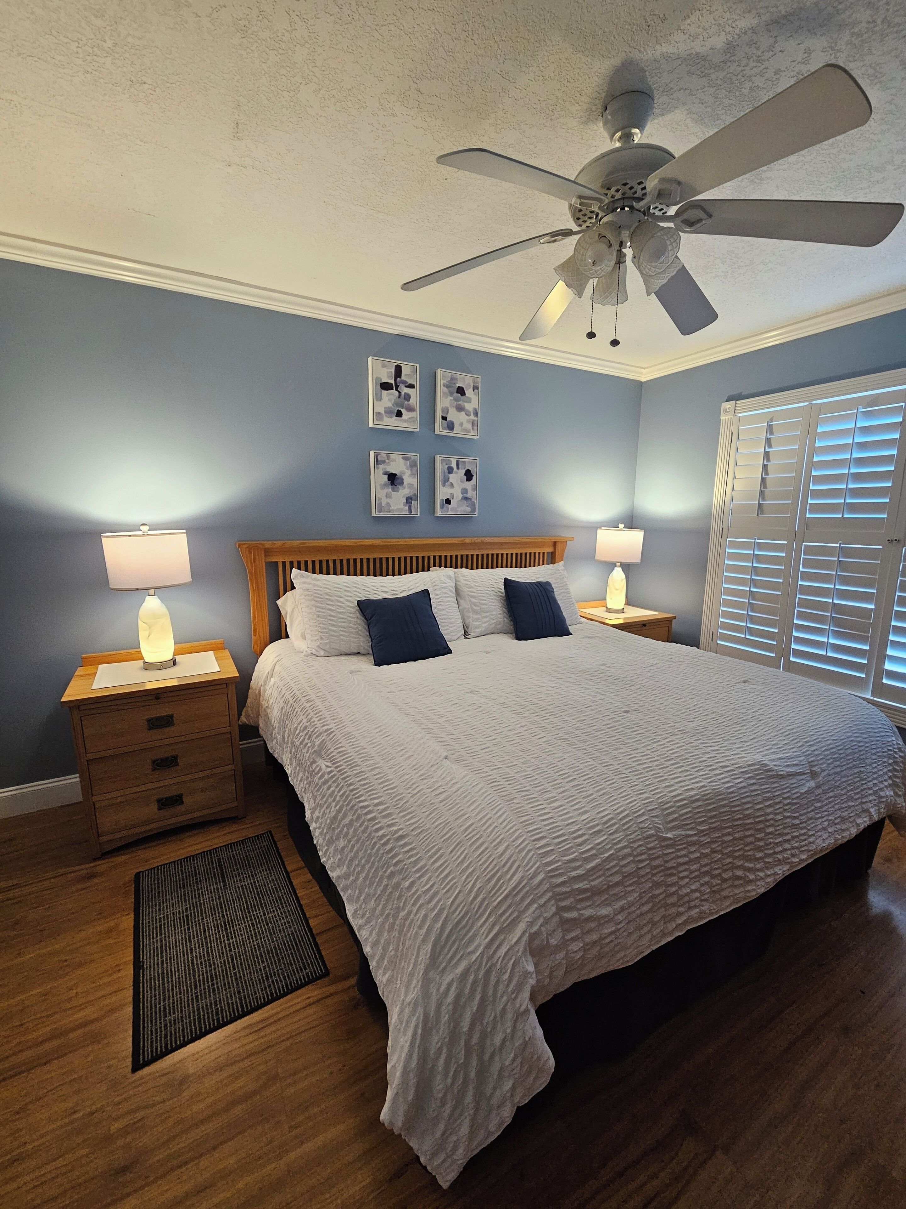 A bedroom with a bed, wooden nightstands, a ceiling fan, and wall art.