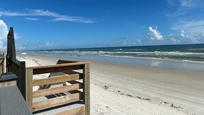Une chaise de plage en bois, placée face à une plage de sable où les vagues déferlent, sous un ciel bleu dégagé.