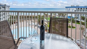 A balcony with a view of the beach, a pool, and a building with a blue roof.