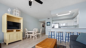 A living room with a TV, wicker table, and a sofa. There is a dining area with chairs and a kitchen in the background.