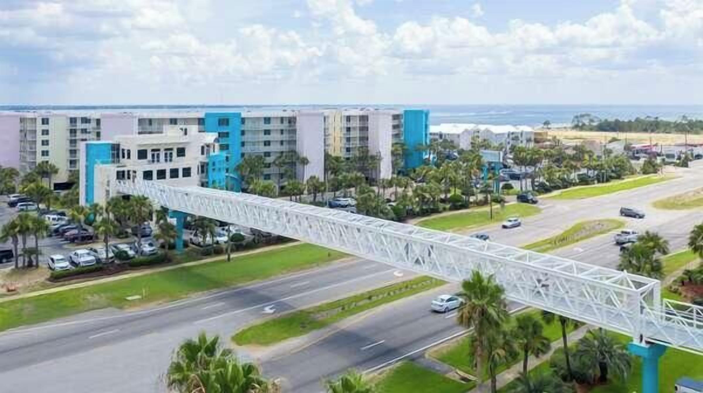 Un hotel de varios pisos con una llamativa estructura de puente blanco en primer plano, un estacionamiento y una vista del océano al fondo.