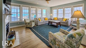 A spacious living room with a large sectional sofa, a coffee table, and a ceiling fan. There are large windows with white frames, a fireplace, and a wicker chair.