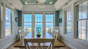 A dining room with a long wooden table, white chairs, and large windows offering a view of the beach.