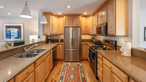 A modern kitchen with granite countertops, stainless steel appliances, wooden cabinets, and a colorful rug. - Wind River 3: Teton Village-Walk to Ski/Lift Sleeps 10- Pool / Hot Tub Access (Teton Village)