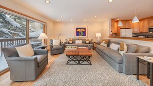 A cozy living room with a fireplace, a large window with a view of snow-covered trees, and a kitchen area with wooden cabinets and pendant lights. - Wind River 3: Teton Village-Walk to Ski/Lift Sleeps 10- Pool / Hot Tub Access (Teton Village)
