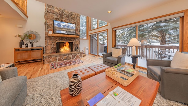 A cozy living room with a stone fireplace, comfortable seating, and large windows offering a view of the outdoors. - Wind River 3: Teton Village-Walk to Ski/Lift Sleeps 10- Pool / Hot Tub Access (Teton Village)