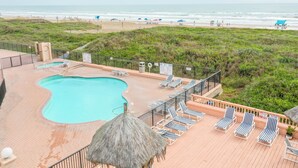 Zona de piscina con sillas de descanso y una sombrilla de paja, con vista a una playa con sombrillas y gente.