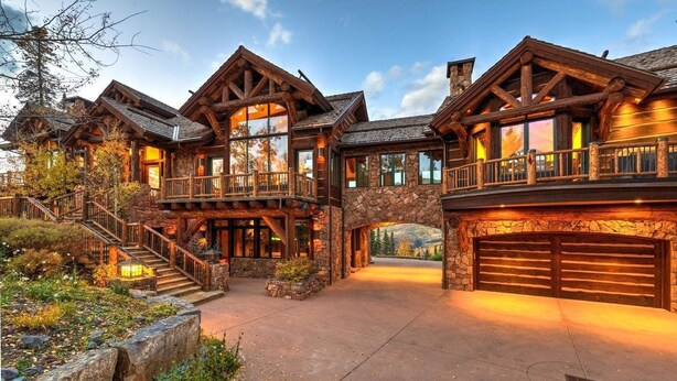A large, multi-story log cabin with a stone foundation, a covered outdoor area, and a stone staircase leading to the entrance.