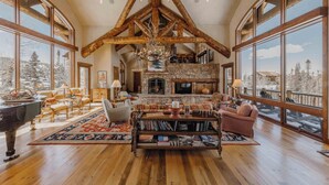 A spacious living room with a grand wooden ceiling, a stone fireplace, and large windows offering a view of snowy trees.