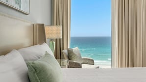 Quarto de hotel com cama, uma cadeira, abajur e vista para o oceano.