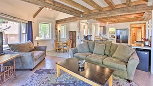 Ein geräumiges Wohnzimmer mit einer großen Couch, einem Couchtisch und einem Essbereich mit Tisch und Stühlen. Das Zimmer zeichnet sich durch freiliegende Holzbalken an der Decke und große Fenster, die für natürliches Licht sorgen, aus.