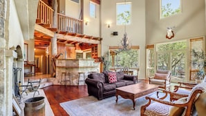A spacious living area with a high ceiling, wooden beams, a fireplace, and a large window overlooking a garden.