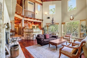A spacious living area with a high ceiling, wooden beams, a fireplace, and a large window overlooking a garden.