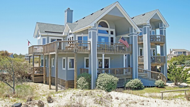 A large, two-story beachfront house with multiple balconies, a covered porch, and a sandy beach in the foreground. - OH1200- 5 BRs with 2 Masters, Private Pool, Hot Tub, Foosball and Volleyball! (Corolla)