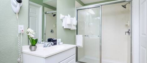 A bathroom with a white vanity, a hairdryer mounted on the wall, a glass shower enclosure, and a potted orchid on the counter.