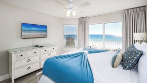 Ein Schlafzimmer mit Bett, Kommode, Deckenventilator, Fernseher und Fenster mit Blick auf den Strand.