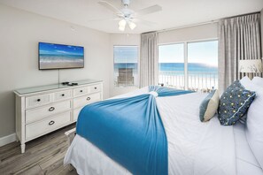A bedroom with a bed, a dresser, a ceiling fan, a TV, and a window with a view of the beach.
