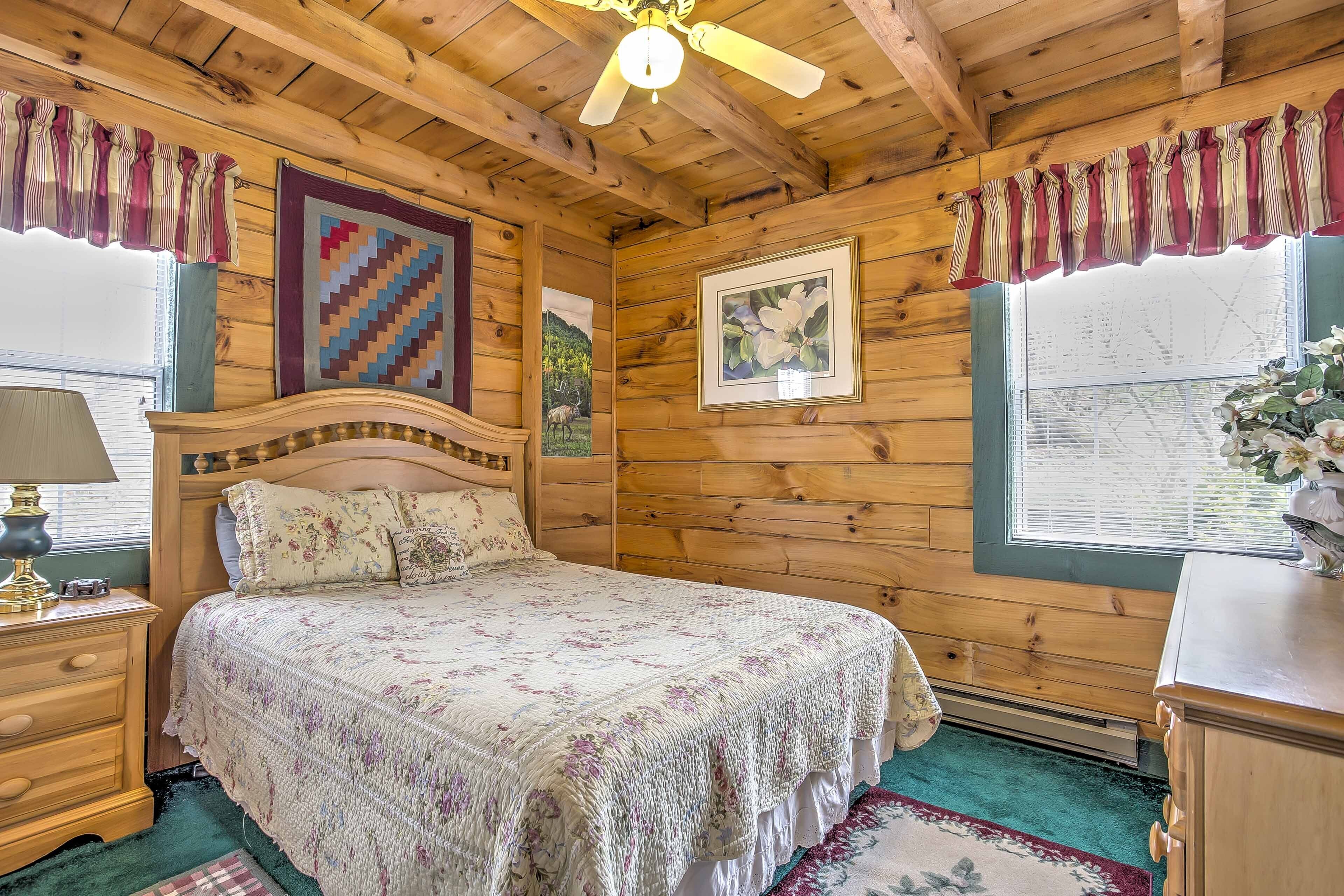 A wooden cabin bedroom with a bed, a nightstand, a window with curtains, and a ceiling fan.