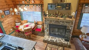 Interior acogedor de una cabaña con chimenea de piedra, paredes de madera, una zona de comedor con mesa y sillas, y un televisor sobre la chimenea.