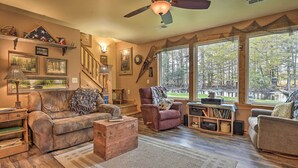 A cozy living room with a wooden floor, a leather sofa, a patterned armchair, and a wooden coffee table. There is a staircase with a wooden railing, a large window with a view of trees, and a ceiling fan. - Secluded Birnamwood Cottage w/ Deck & River Views! (Birnamwood)