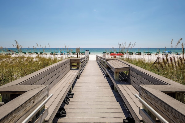 A wooden boardwalk leading to a beach with umbrellas and beachgoers.