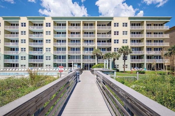 A multi-story hotel with balconies, a pool, and a wooden boardwalk leading to the entrance.