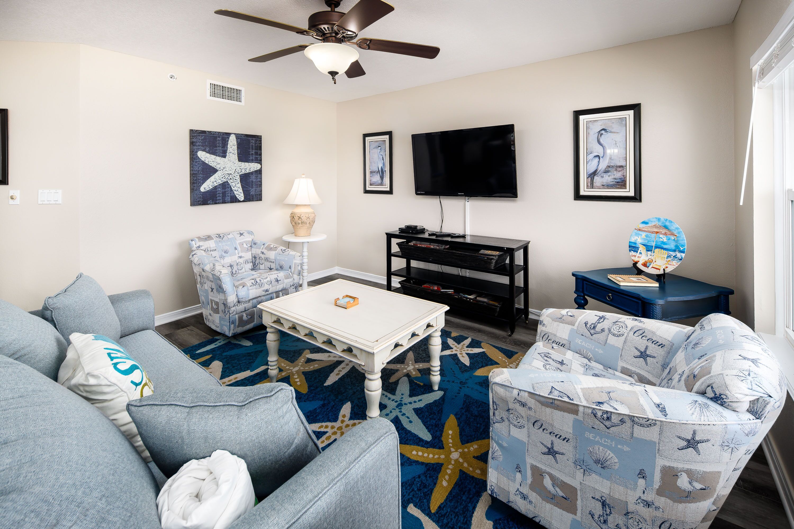 A living room with a ceiling fan, a flat-screen TV, a coffee table, and a patterned rug.