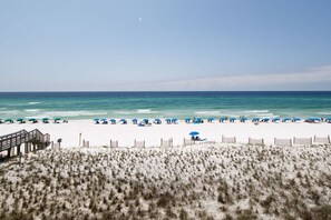 Une plage de sable blanc, parsemée de parasols bleus, et une promenade en bois qui mène à l’eau.