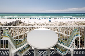 A balcony with a white table and striped chairs overlooking a beach with blue umbrellas.