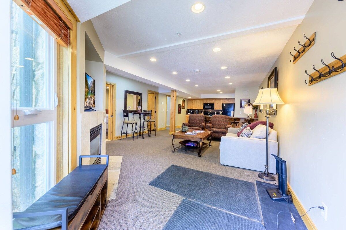 A spacious living area with a sofa, a coffee table, and a bench. There is a kitchen area in the background with a dining table and chairs.