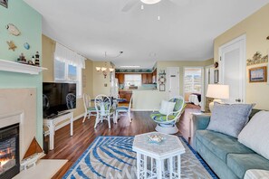 A living room with a fireplace, a dining area with white chairs, a kitchen with wooden cabinets, and a view of the sea through the windows.