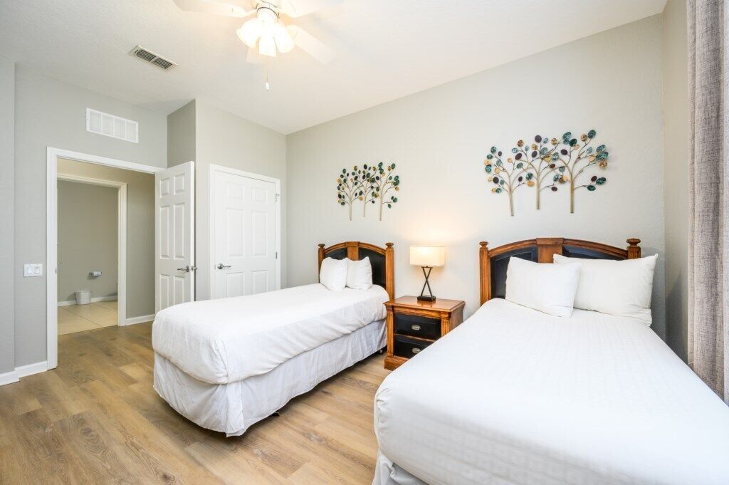 Une chambre d’hôtel avec deux lits simples, une table de chevet en bois avec une lampe et un ventilateur de plafond.