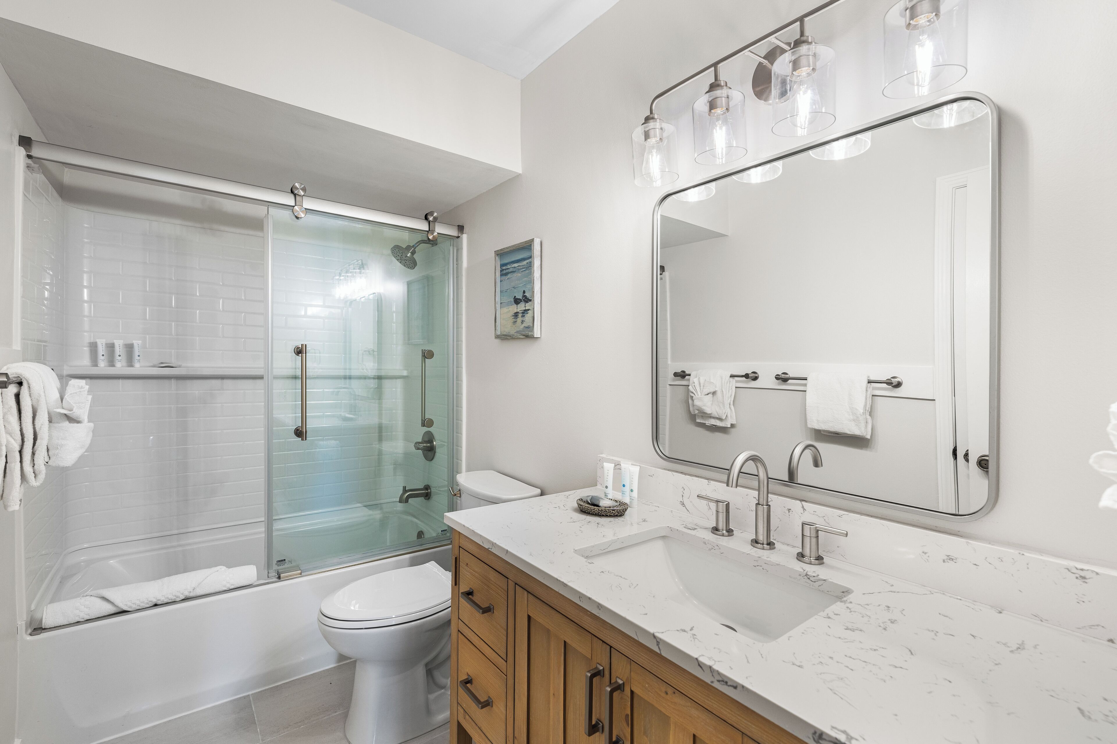 A modern bathroom with a glass-enclosed shower, a white toilet, a sink with a marble countertop, wooden cabinets, and a large mirror with lights.