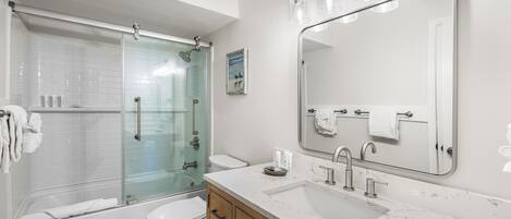 A modern bathroom with a glass-enclosed shower, a white toilet, a sink with a marble countertop, wooden cabinets, and a large mirror with lights.